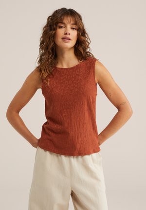 Femme aux cheveux bouclés portant un haut sans manches couleur rouille et un pantalon beige clair, debout les mains sur les hanches contre un fond neutre.