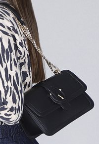 Donna che indossa una borsa a tracolla in pelle nera con una cintura e dettagli in oro, abbinata a una maglietta a maniche lunghe con motivo.