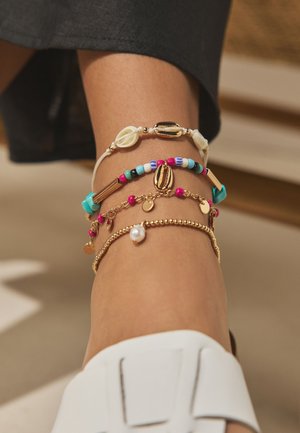 Ankle with four colorful beaded and shell anklets paired with white slip-on shoe and black pants.
