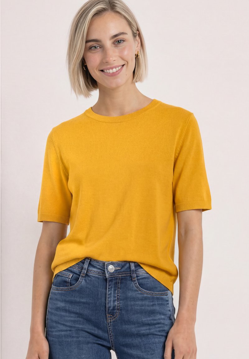 Femme souriante aux cheveux blonds courts, vêtue d'un haut jaune moutarde à manches courtes et d'un jean bleu, sur un fond clair uni.