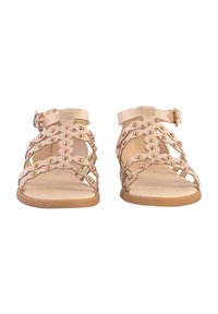 Sandalias beige claro con tiras entrelazadas, adornadas con tachuelas doradas y una correa para el tobillo con hebilla. Suela plana con superficie interior texturizada.