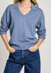 Femme portant un pull col V bleu clair avec col et un jean bleu foncé, mains dans les poches, devant un fond uni.