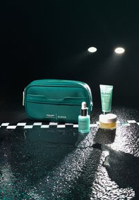 Trousse de maquillage teal avec fermeture éclair, contenant un sérum teal, une crème dans un pot et un tube ; tous étiquetés "ELEMIS". Gouttes d'eau sur la surface.