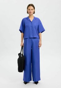 Mėlyna, trumpomis rankovėmis marškinėliai su krūtinės kišene, derinami su plačiomis mėlynomis kelnėmis. Modelis laiko juodą pintą rankinę. Paprastas, modernus dizainas.