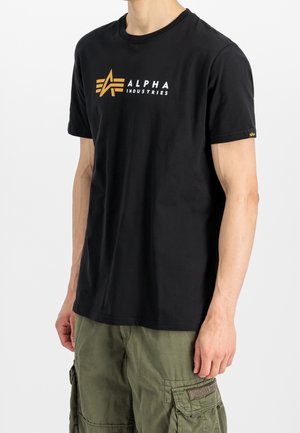 Mężczyzna w czarnej koszulce Alpha Industries z pomarańczowym logo i oliwkowozielonych spodniach cargo na jasnym, jednolitym tle.