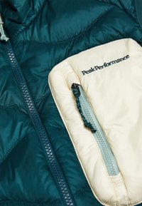 Turquoise doorgestikte jas met een beige zak met rits, voorzien van een ritstrekker en "Peak Performance" geborduurd in zwarte draad.