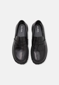 Svarta läderslip-on skor med en slät yta, runda tår, och ett dekorativt rem över fotryggen. Gummisula för bättre grepp.
