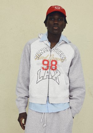 Giovane uomo che indossa una felpa con cappuccio grigia con zip e testo "NINE4STEEL 98 LAX", camicia azzurro chiaro sotto, pantaloni della tuta grigi e un berretto rosso.