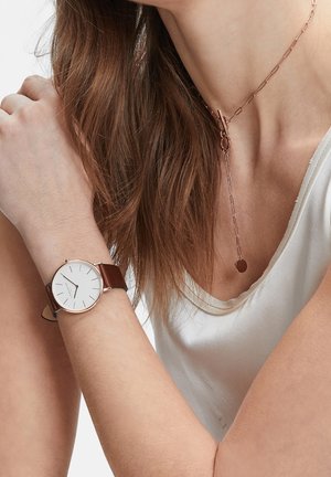 Femme portant un haut blanc simple sans manches, un collier chaîne en or rose avec un pendentif circulaire, et une montre-bracelet avec un bracelet en cuir marron.