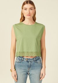 Top corto in cotone verde con bordi in pizzo scallopato all'orlo, dotato di scollatura rotonda e design senza maniche, abbinato a jeans blu chiaro.