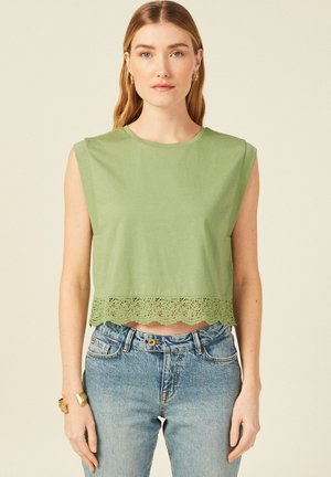 Crop top en coton vert avec un ourlet en dentelle festonné, comportant un col rond et un design sans manches, associé à un jean bleu clair.