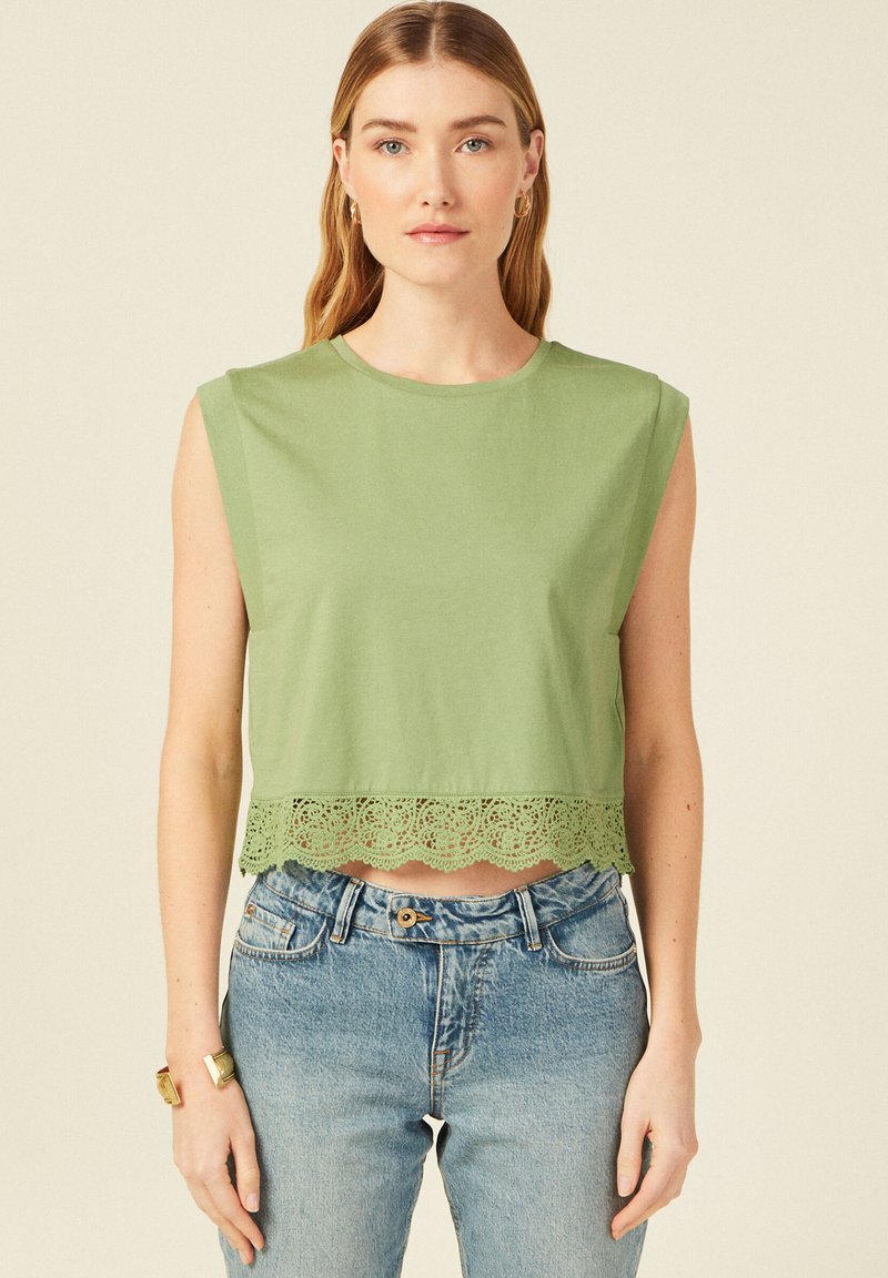 Top corto in cotone verde con bordi in pizzo scallopato all'orlo, dotato di scollatura rotonda e design senza maniche, abbinato a jeans blu chiaro.