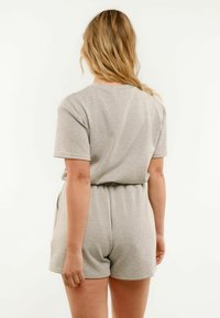 Frau mit blondem Haar, die ein hellgraues kurzärmeliges Oberteil und passende Shorts mit elastischem Bund trägt, steht mit dem Rücken zur Kamera vor einem schlichten Hintergrund.