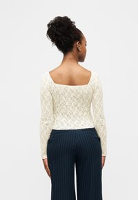 Γυναίκα με σγουρά μαλλιά δεμένα ψηλά, φοράει λευκό μακρυμάνικο τοπ από δαντέλα και μπλε σκούρο παντελόνι με κάθετες ρίγες, στέκεται πλάτη σε απλό φόντο.