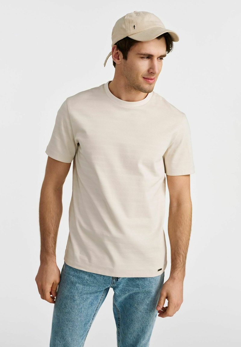 Beżowa koszulka z krótkim rękawem, z okrągłym dekoltem, wykonana z miękkiego materiału. Zestawiona z jasnoniebieskimi dżinsami i jasnobeżową czapką.