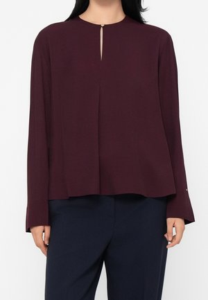 Kvinde iført en mørk burgunderfarvet bluse med lange ærmer og en lille nøglehul foran samt marineblå bukser mod en ensfarvet baggrund.