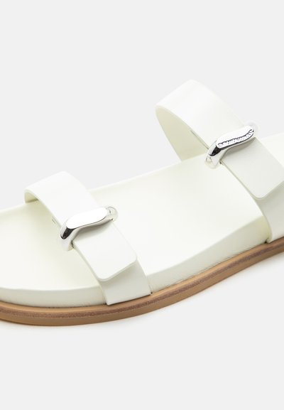 Λευκό σανδάλι με δύο ρυθμιζόμενους ιμάντες που διαθέτουν ασημένιες αγκράφες και ανοιχτό καφέ σόλα, εμφανίζεται σε απλό φόντο.