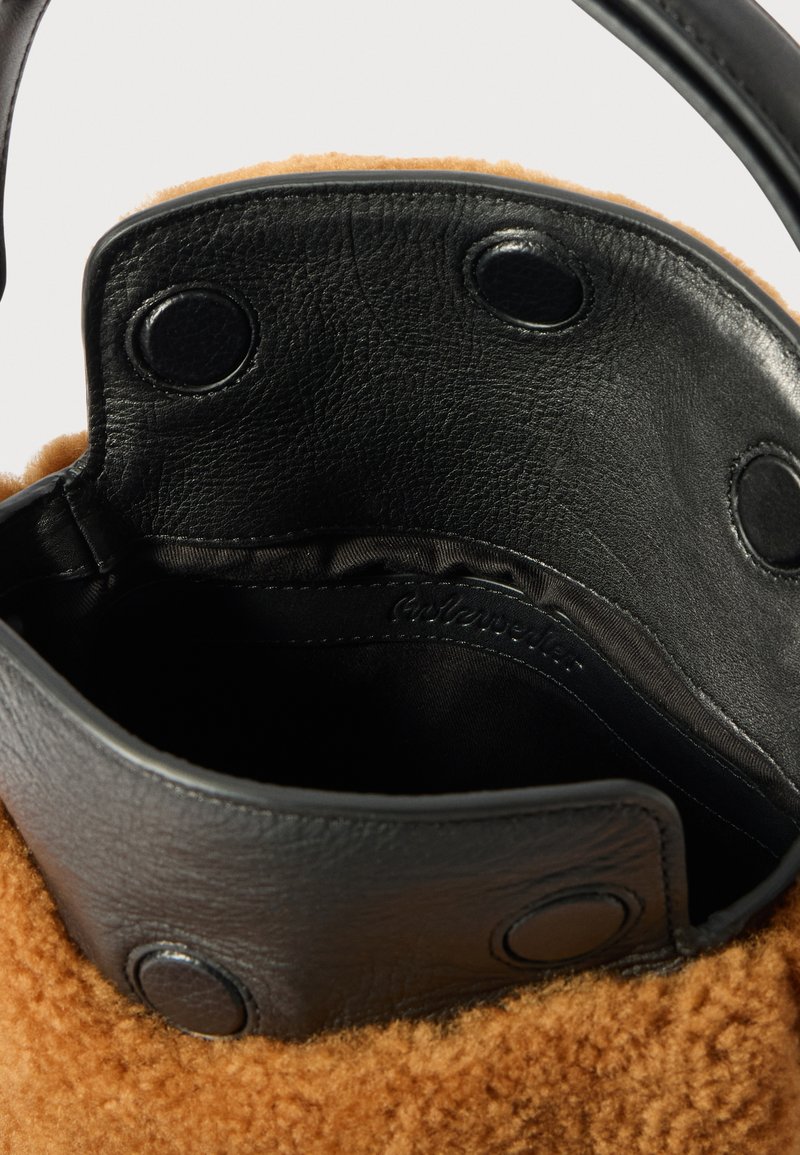 Primo piano di una borsa in shearling marrone con pattina in pelle nera fissata da tre bottoni a pressione neri e fodera interna nera liscia visibile.