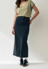 Mørk denim maxi-nederdel med lige snit og frynset kant, parret med en lys grøn t-shirt med v-udskæring og sorte sneakers med hvide detaljer.