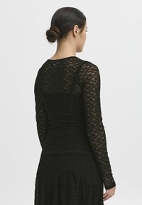 Top lungo in pizzo nero con maniche lunghe, design aderente, motivo floreale testurizzato e scollo rotondo. Il tessuto è semitrasparente.