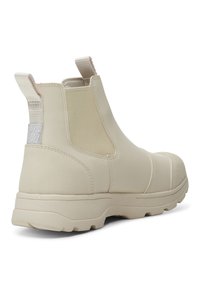 Botín de tobillo de goma beige con paneles laterales elásticos, parte trasera texturizada y suela plana. Presenta unaasa de tirón para facilitar el uso y un acabado liso y mate.
