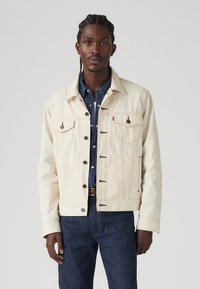 Beige denim jack met oranje stiksels, knoopsluiting aan de voorkant en twee borstzakken. Draagt het over een blauwe shirt; model staat voor een vlakke achtergrond.