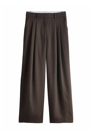 Hand justiert dunkelbraune maßgeschneiderte Hose mit passender Weste, zeigt Bauch und Goldring an der linken Hand.