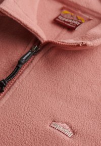Lichtroze fleece halve-rits trui met een gestructureerde afwerking, voorzien van een logopatch en een zwarte ritssluiting met een stoffen trekkoord.