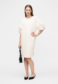 Ženska v belih oblačilih, ki segajo do kolen, z napuhnjeniimi rokavi, drži črno torbico in nosi črne mule z ostrim nosom, stoji na belem ozadju.