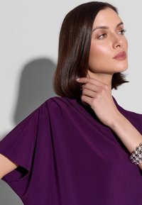 Fioletowa bluzka z krótkimi rękawami, miękki materiał, zdobiona marszczeniem na ramionach. Srebrna bransoletka na nadgarstku. Proste tło.