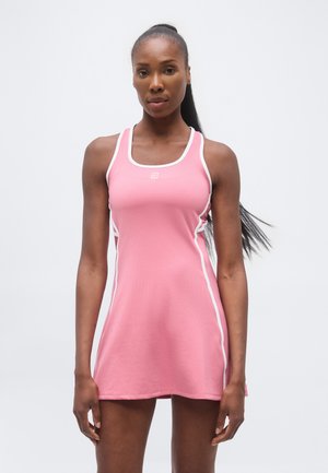 Femeie cu păr lung care poartă o rochie sport roz fără mâneci cu margine albă, stând în fața unui fundal simplu și deschis.