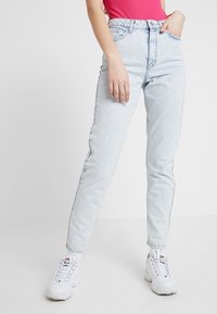 Vaaleansiniset korkeavyötäröiset farkut suoranlahkeisella leikkauksella, näkyvillä saumoilla ja viidellä taskulla, yhdistettynä valkoisiin tennareihin.