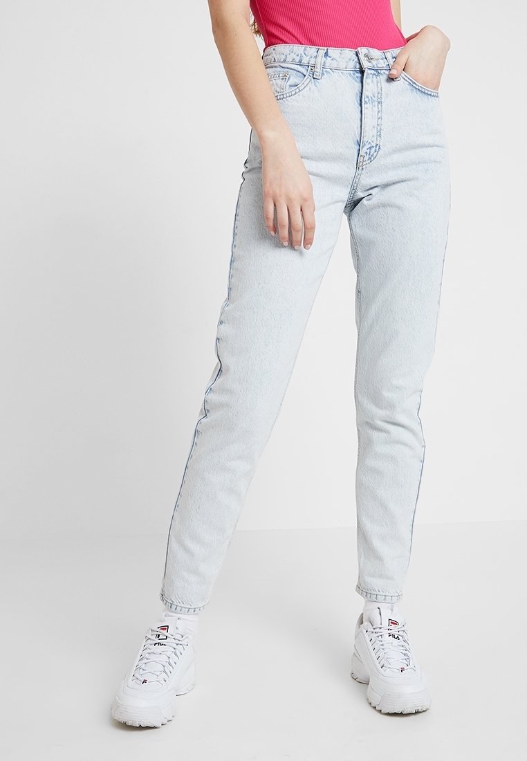 Vaaleansiniset korkeavyötäröiset farkut suoranlahkeisella leikkauksella, näkyvillä saumoilla ja viidellä taskulla, yhdistettynä valkoisiin tennareihin.