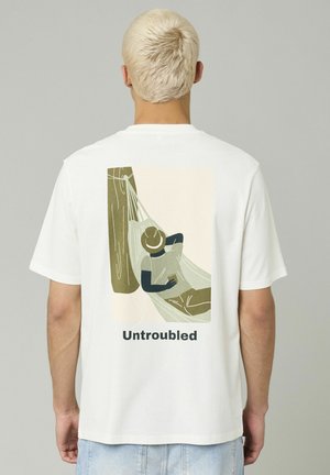 T-shirt en coton blanc avec une illustration imprimée d'une personne se relaxant dans un hamac, avec le mot "Sans souci" en dessous de l'image.