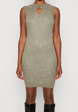 Ärmelloses olivgrünes Strickkleid mit geometrischem Muster, Ausschnitt in Tropfenform und goldfarbenen Knopfdetaills auf den Schultern, kombiniert mit schwarzen Stiefeln.