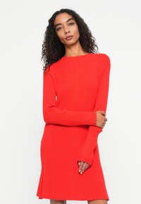 Abito rosso in maglia a costine con maniche lunghe, scollatura a barca e orlo svasato. Il tessuto presenta linee di texture diagonali per un dettaglio aggiuntivo.