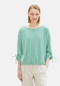 Türkiser gestreifter Pullover mit langen Ärmeln und gebundenen Bündchen, im lässigen Schnitt und mit strukturiertem Stoff. Kombiniert mit hellen Hosen.