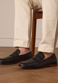 Zwarte leren instappers met een geprofileerde afwerking en een ronde neus, voorzien van een brede band over de wreef. Gepaard met beige broeken.
