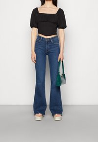 Svart kort topp med puffärmar, blå utsvängda jeans, vita plattformssandaler och en grön handväska. Slät textur och skräddarsydd passform.