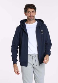 Sweat à capuche zippé marine en tissu mélangé coton. Il présente une poche frontale, des poignets côtelés et un patch logo sur la poitrine gauche. Associé avec un pantalon de jogging gris.