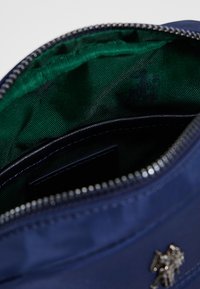 Laivastonsininen kangaskassi hopeanvärisellä vetoketjulla ja vihreällä vuorauksella sisäpuolella. Edessä pieni koristeellinen tunnus.