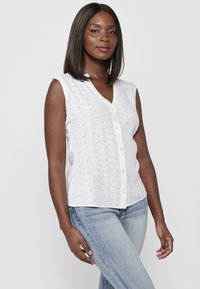Blouse blanche sans manches avec un design en œillets texturés, décolleté en V et fermeture à boutons à l'avant ; associée à un jean bleu clair.