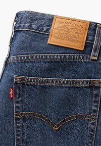 Poche arrière d'un jean bleu en denim Levi's avec couture orange, étiquette rouge Levi's et patch en cuir beige sur la ceinture.