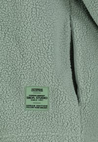 Fleecejacke in Hellgrün mit einer strukturierten Oberfläche, versehen mit einem rechteckigen grünen Etikett, das Marken- und Textinformationen enthält.
