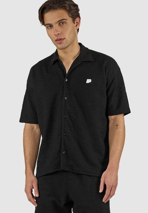 Joven con cabello castaño que lleva una camisa negra de manga corta con textura y botones, y pantalones a juego, mostrando un pequeño tatuaje de cruz en el antebrazo izquierdo.