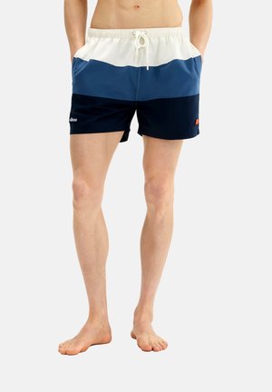 Persona con las piernas desnudas que lleva puestos unos pantalones cortos de baño con bloques de color azul, navy y blanco, con cordón y bolsillos laterales, de pie sobre un fondo liso.