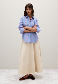 Gestreiftes, blaues und rosafarbenes Hemd mit umgeschlagenen Ärmeln, kombiniert mit einem cremefarbenen Maxirock und braunen, flachen Espadrilles. Die Textur ist glatt.