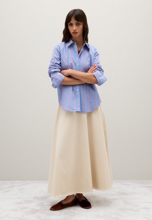 Camicia button-up a righe blu e rosa con maniche arrotolate, abbinata a una gonna maxi color crema e espadrillas piatte marroni. La texture è liscia.