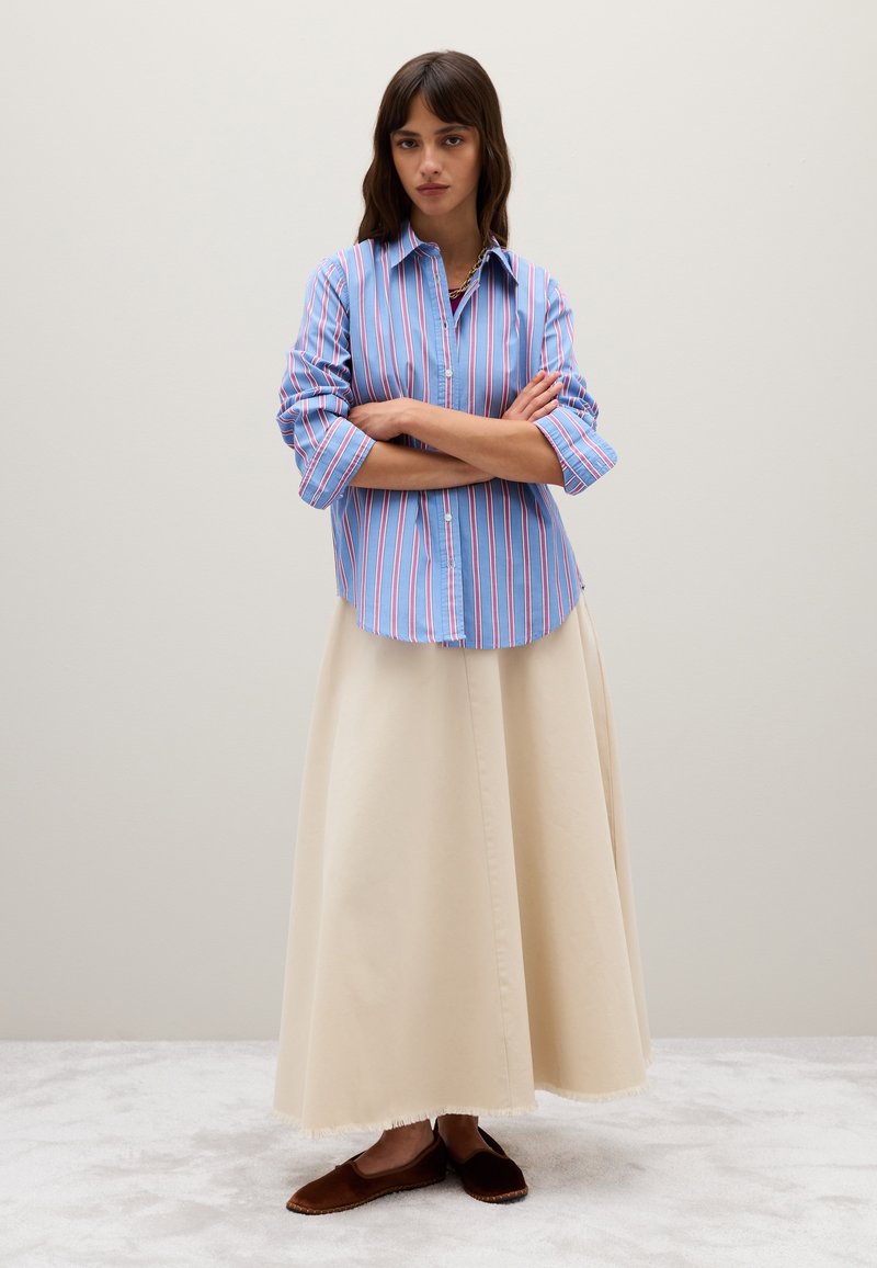 Gestreiftes, blaues und rosafarbenes Hemd mit umgeschlagenen Ärmeln, kombiniert mit einem cremefarbenen Maxirock und braunen, flachen Espadrilles. Die Textur ist glatt.