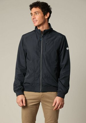 Navy blauwe jack met ritssluiting en hoge kraag, elastische manchetten en een borstzak. Gemaakt van lichtgewicht materiaal met een gladde textuur.